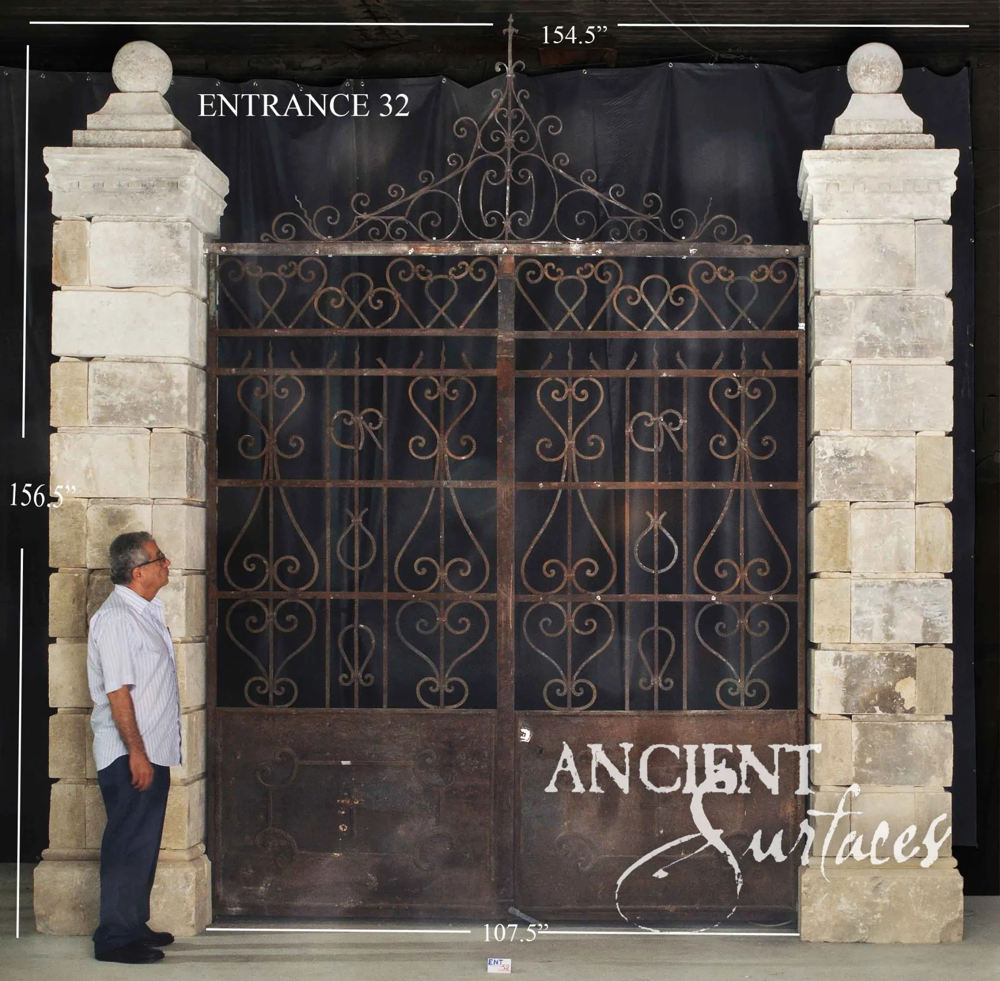 Monumental reclaimed limestone gate piers from Ancient Surfaces topped with carved stone spheres, flanking a wrought iron estate gate with scrolled cresting, a man standing alongside for scale