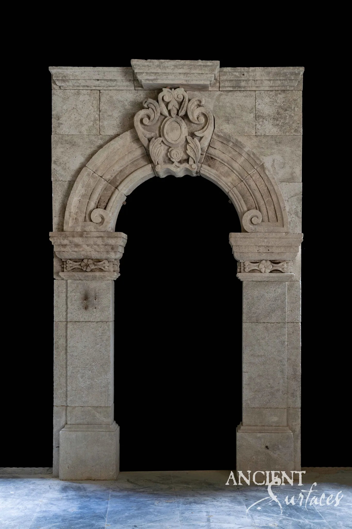Reclaimed antique limestone entryway from Ancient Surfaces with a carved shield keystone and a flat capped lintel above moulded impost capitals, photographed on a marble floor
