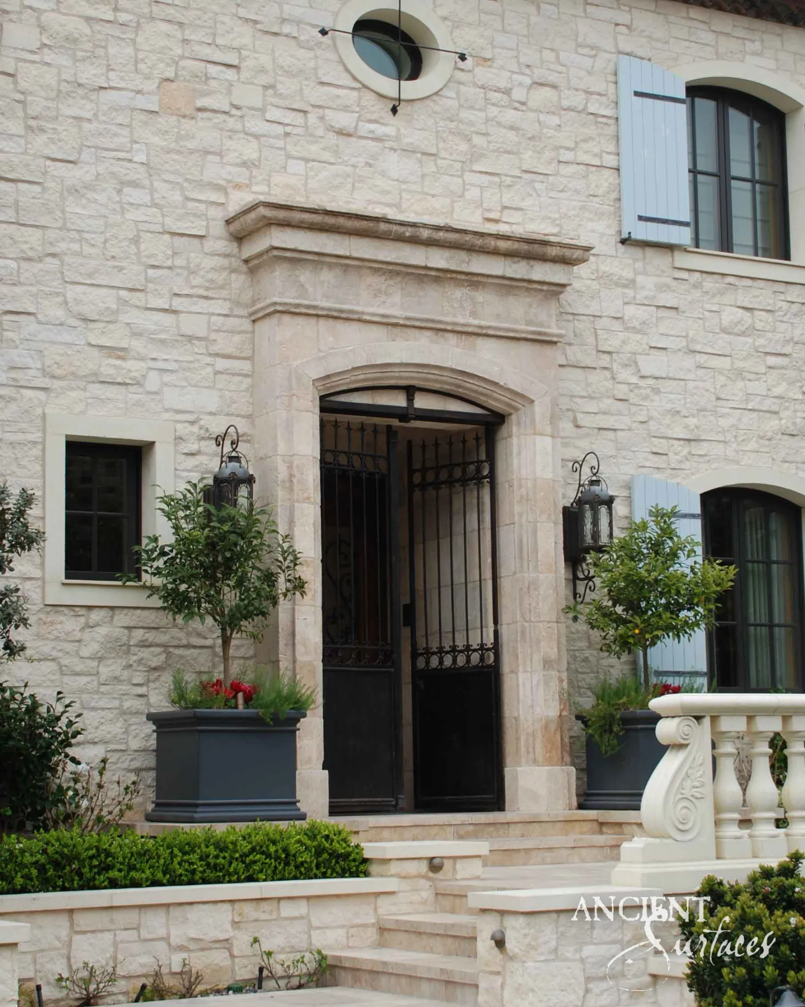 Reclaimed antique limestone entryway from Ancient Surfaces with a segmental arched surround and iron gate, set within a fully stone clad French facade beneath an oculus window